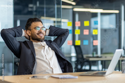 A businessman wearing glasses smiles and relaxes in his office, with his hands behind his head.