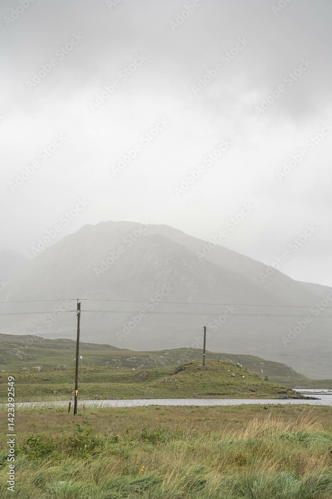 Fototapeta premium Scenic lake in the Connemara region of Ireland, surrounded by lush green hills, cloudy skies, and a traditional stone wall in the foreground.