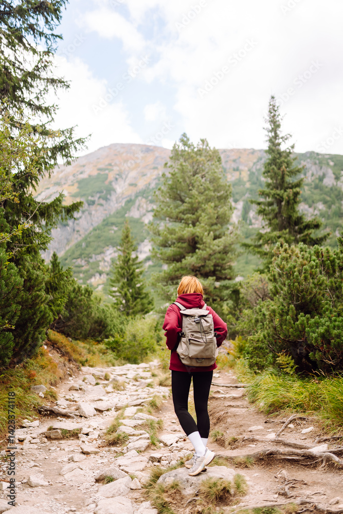 Naklejka premium Woman traveler with backpack walking along rocky hiking trail in mountains. Happy woman enjoying nature and feeling freedom in open air. Hiking, adventure concept.