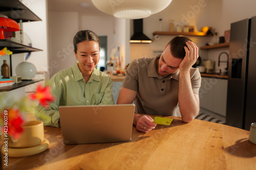 Happy couple shopping online, using a laptop and credit card, enjoying quality time together in their stylish, modern kitchen filled with comfort and convenience