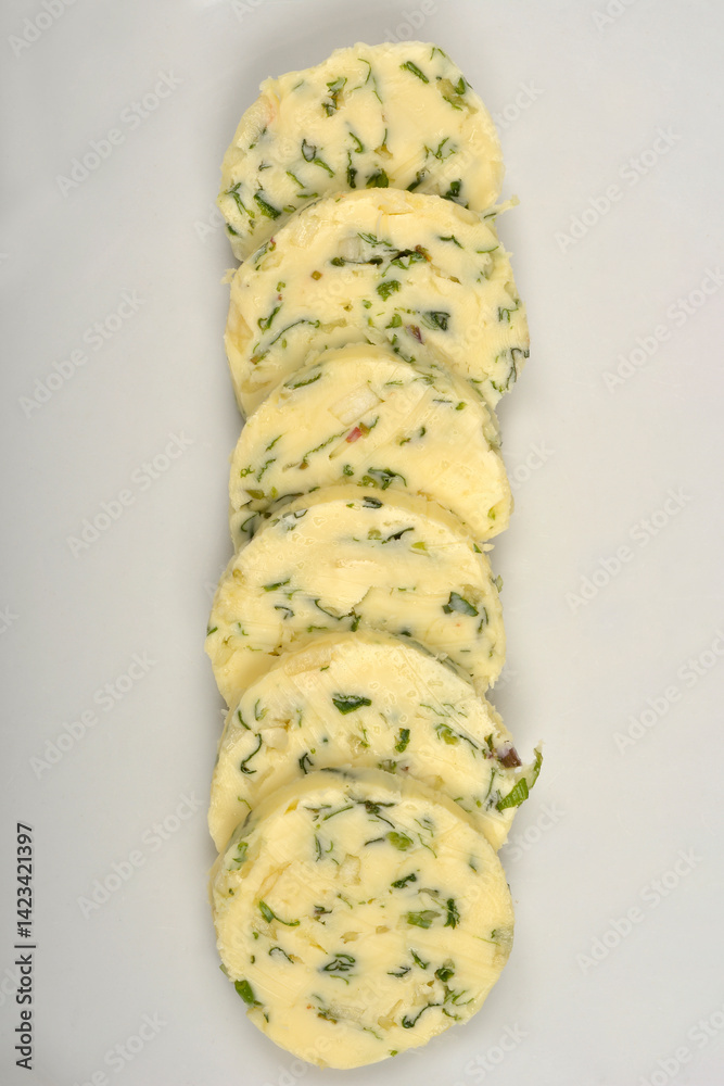 Slices of pale yellow butter with green herbs and garlic are arranged vertically on a white plate, captured from a top-down perspective