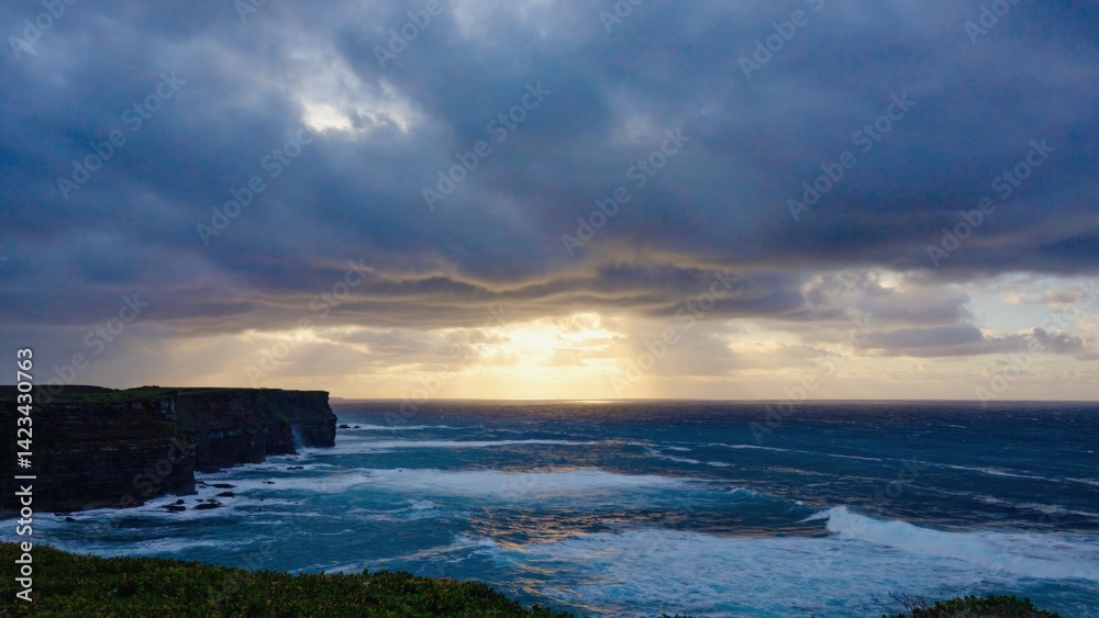 Serene view of sun setting over ocean, with sky full of clouds and cliff's edge in background. Serene view of sun setting over ocean, with sky full of clouds and cliff's edge in background.