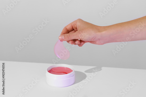 woman’s hands taking pink hydrogel patch from a white jar on a white background. hydrate, soothe, and nourish, diminishing dark circles and fine lines for fresh, bright under-eyes concept