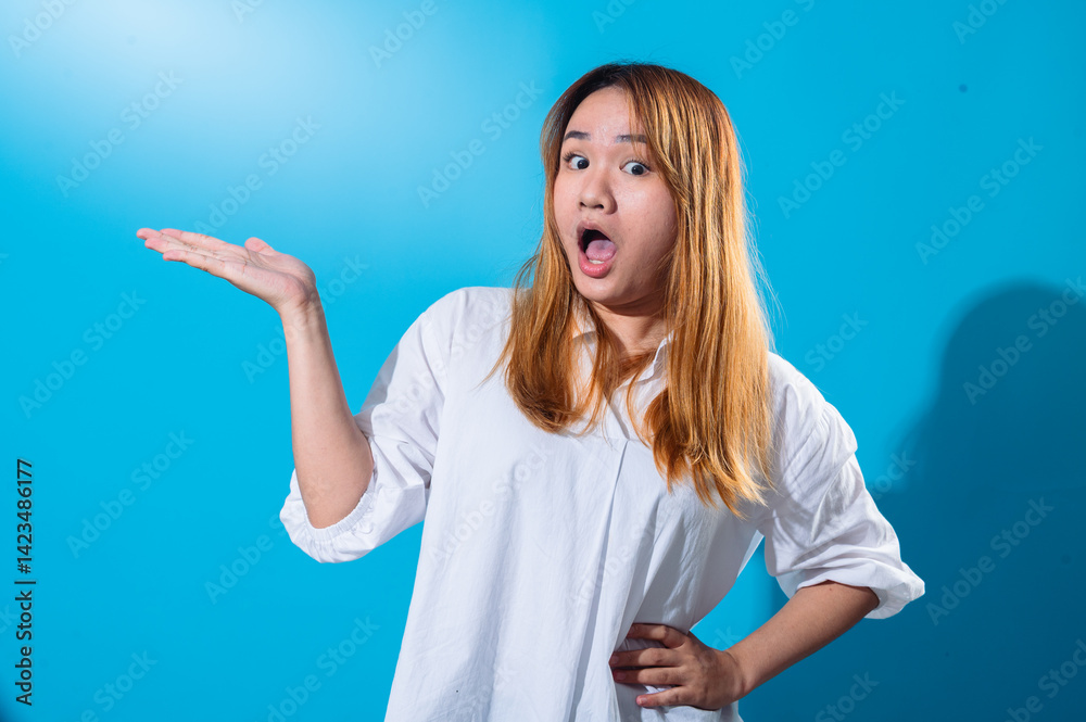 Fototapeta premium Asian woman with long straight hair in white shirt smiling while holding one hand open to the side as if presenting or showing something, standing confidently against a solid blue studio background