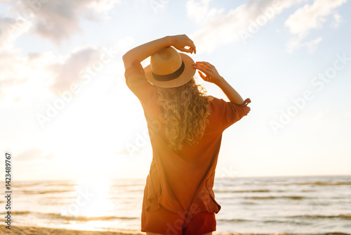 Back view of a young woman with curly hair feels free on the seashore. Young woman walks on the beach at sunset. Concept of relaxation, enjoyment, freedom.