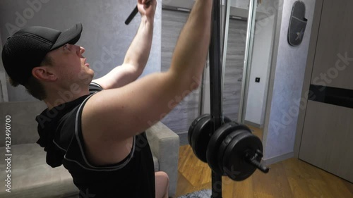 Young and handsome Athlete sportsman pumps up his back muscle on a simulator with a heavy weight. Workout at home in modern apartments. Slow motion. Close-up.