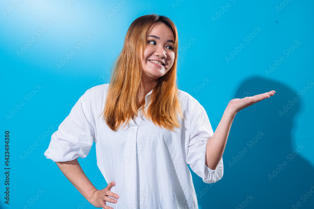 Fototapeta premium Asian woman with long straight hair in white shirt smiling while holding one hand open to the side as if presenting or showing something, standing confidently against a solid blue studio background