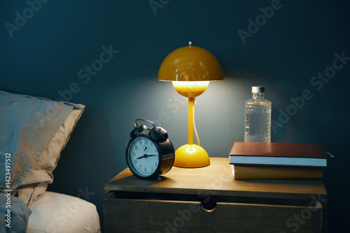 Papier peint Cozy bedroom nightstand setup showcasing a yellow lamp, an alarm clock, books, a