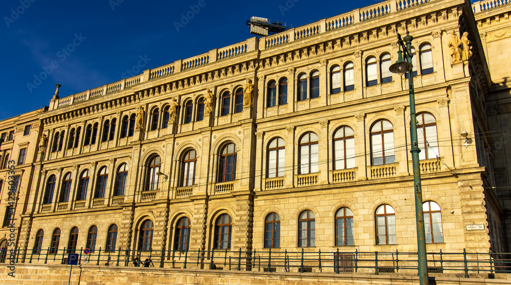 Facade of historic neoclassical building with arched windows and sculptures in Budapest, Hungary, illuminated by warm sunlight under clear blue sky Facade of historic neoclassical building with arched windows and sculptures in Budapest, Hungary, illuminated by warm sunlight under clear blue sky