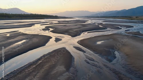 Aerial view of a wide, braided river flowing through a flat valley at sunrise, with distant mountains silhouetted against the soft, golden sky and trees alongside the water.
