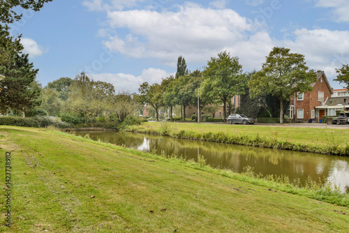 A tranquil scene featuring a gently flowing waterway bordered by vibrant green grass and trees, with charming houses in the background under a clear blue sky.