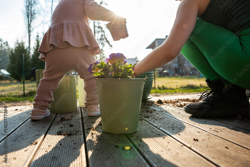 A child in pink holds a container while an adult in green leggings tends plants on a wooden deck. Potted purple flowers and sunlight enhance the scene.