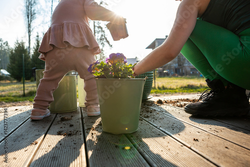 Obraz na płótnie A child in pink holds a container while an adult in green leggings tends plants on a wooden deck. Potted purple flowers and sunlight enhance the scene. w salonie