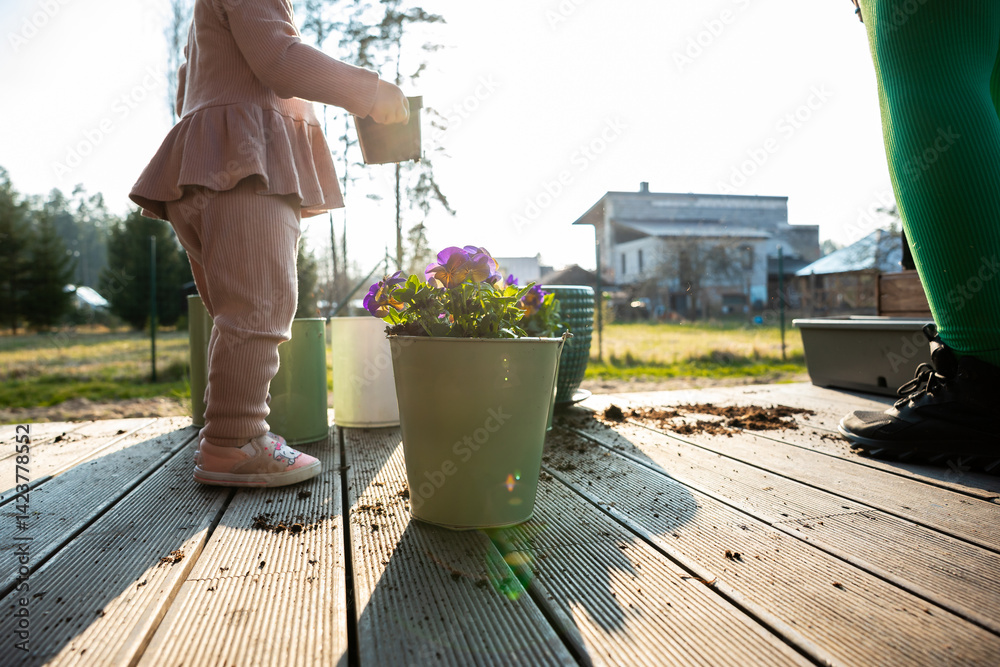 A child in pink stands on a wooden deck holding a gardening tool near a green pot with purple flowers. A modern house and open field are in the background.