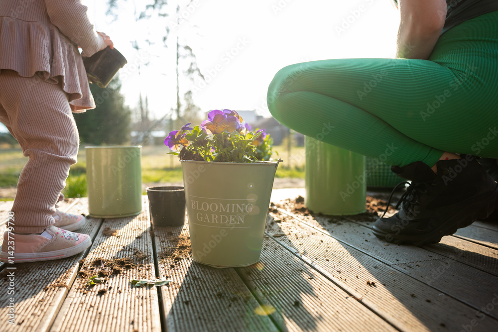 A child and adult engage in gardening on a wooden deck with a green pot labeled BLOOMING GARDEN, vibrant purple flowers, and scattered tools under sunlight.