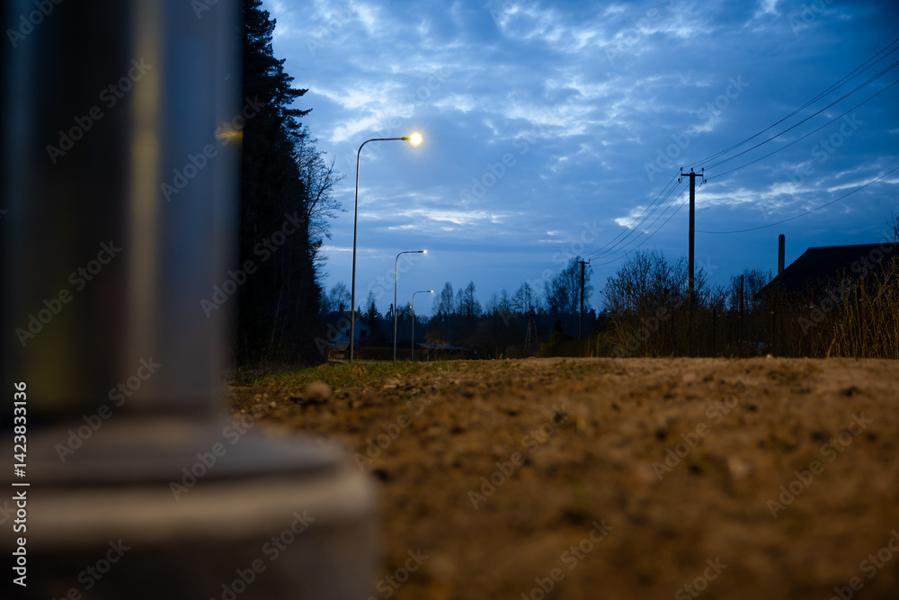 A dirt pathway lit by streetlights under a deep blue evening sky, with power lines, a utility pole, tree silhouettes, and a blurred metallic object in view.