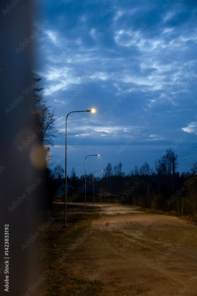 A quiet dirt road lit by streetlights under a dim evening sky with scattered clouds. Sparse trees and distant houses create a rural atmosphere.