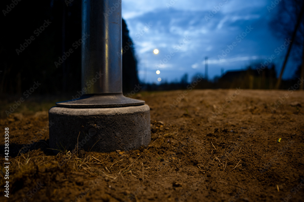 Low angle view of a metallic pole's base on a dirt pathway, with concrete foundation, faint streetlights, trees, and a partially clouded blue sky at dusk.