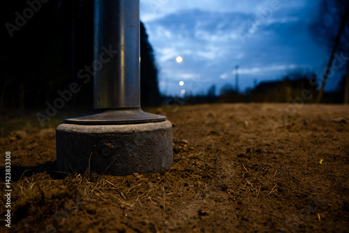 Obraz na płótnie Low angle view of a metallic pole's base on a dirt pathway, with concrete foundation, faint streetlights, trees, and a partially clouded blue sky at dusk. w salonie