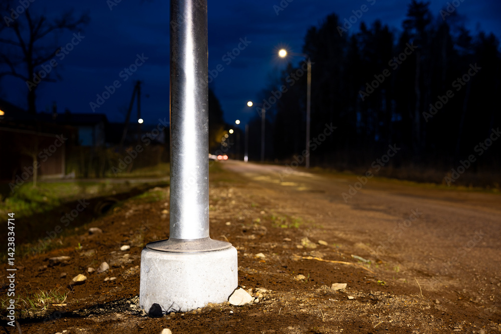 A metal streetlight pole anchored in concrete stands along a dimly lit road at night, with scattered rocks, dirt, and faint outlines of trees visible.