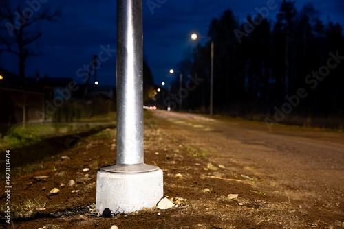 Obraz na płótnie A metal streetlight pole anchored in concrete stands along a dimly lit road at night, with scattered rocks, dirt, and faint outlines of trees visible. w salonie