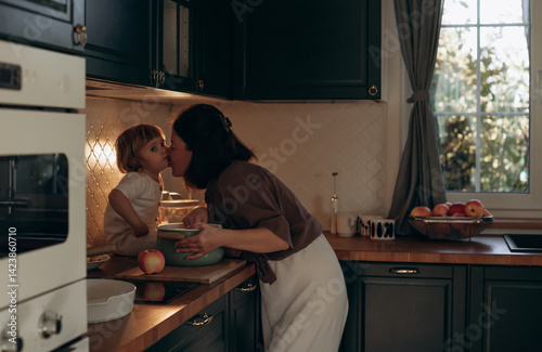 A mother kisses her young daughter in a warm kitchen filled with natural light. They are interacting near a bowl while apples are visible on the countertop, creating a homey atmosphere