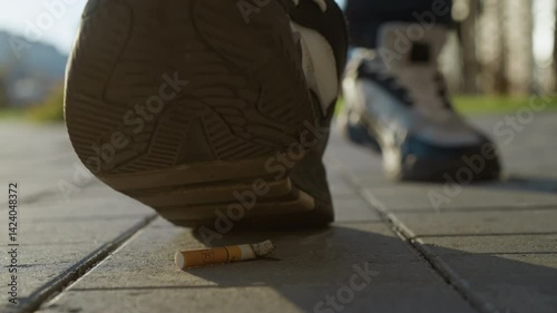 close up of burning cigarette lying on paved sidewalk with smoke swirling upward as person walks toward it and prepares to crush it underfoot
