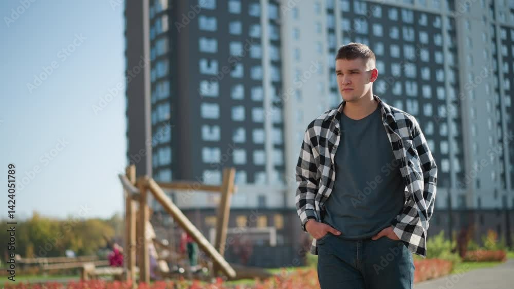 student in walks with hands in pockets while looking around thoughtfully during sunny day in modern residential area with tall buildings and blurred children playing near wooden playground structure