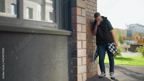 Wallpaper Mural young male wearing puffer vest and checkered shirt tied around waist leans against brick wall while inhaling vape device and coughing on urban street with modern buildings in bright natural light Torontodigital.ca
