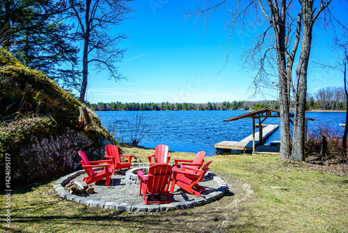 Cottage escape: fire pit with circle of red wooden arm chairs with lake dock and blue sky in background shot in cottage country in early spring