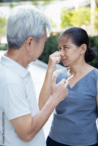 Heavy smoker with bad breath, old Asian woman pinching her nose due to bad breath, foul smell, stinky unpleasant annoying odor from the mouth of smoking man