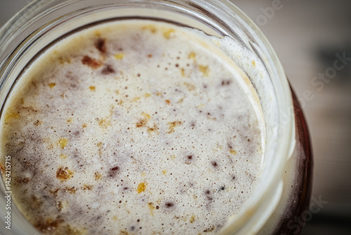 Photos A close-up of the surface of fermenting honey with a frothy texture and dark spots in a glass jar, showing active microbial activity