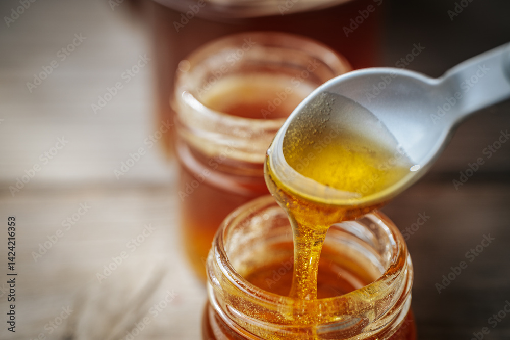 A close-up of golden honey slowly dripping from a plastic spoon into a glass jar, with more jars in the background softly blurred on a rustic wooden surface. A close-up of golden honey slowly dripping from a plastic spoon into a glass jar, with more jars in the background softly blurred on a rustic wooden surface.
