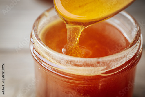 A close-up of golden honey dripping from a wooden spoon into a glass jar, showcasing its rich, smooth texture and natural color. Warm tones dominate the composition.