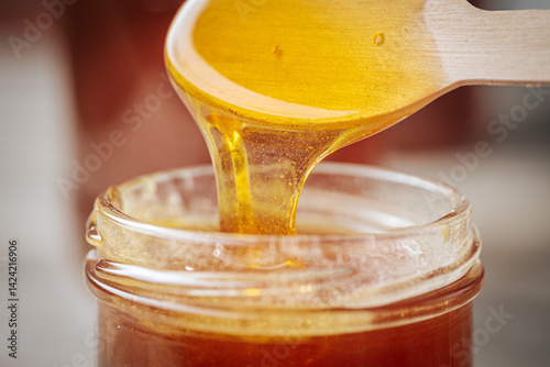 Fotografija A close-up of golden honey dripping from a wooden spoon into a glass jar, showcasing its rich, smooth texture and natural color