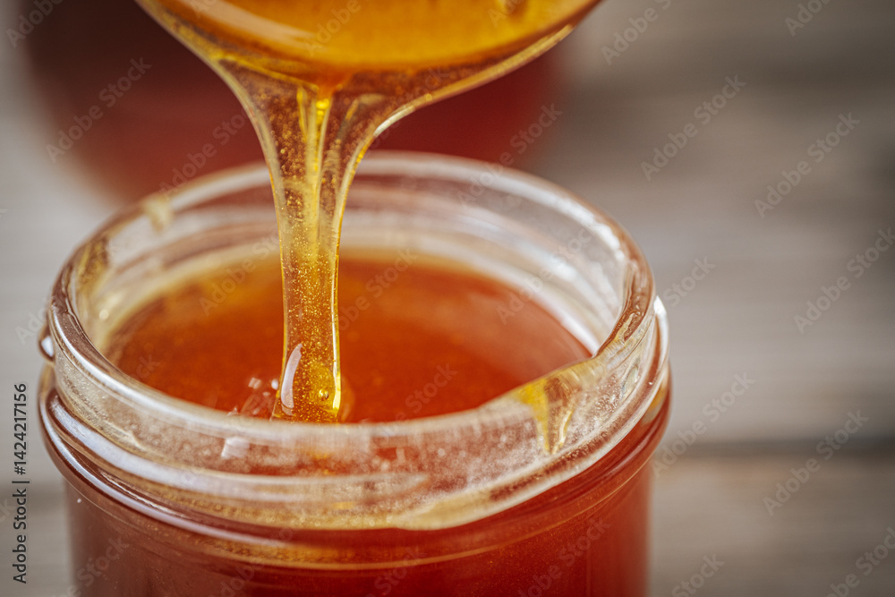A close-up of golden honey dripping from a wooden spoon into a glass jar, showcasing its rich, smooth texture and natural color. Warm tones dominate the composition. A close-up of golden honey dripping from a wooden spoon into a glass jar, showcasing its rich, smooth texture and natural color. Warm tones dominate the composition.