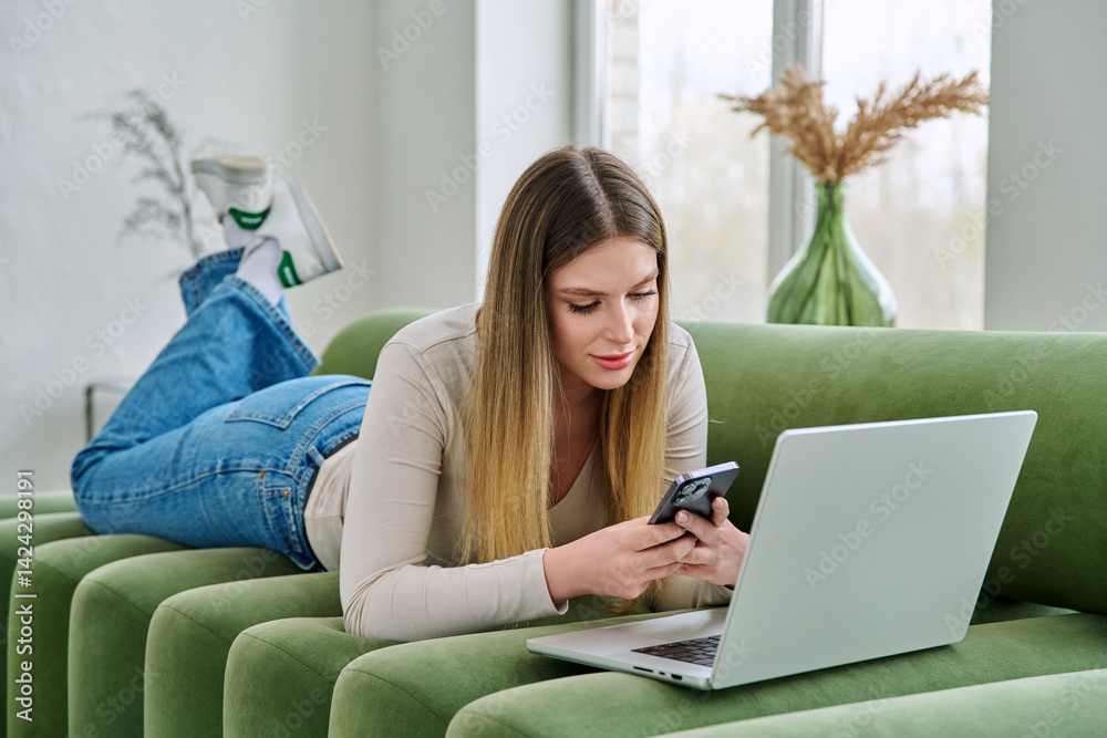 Young serious woman lying on couch with laptop, using smartphone