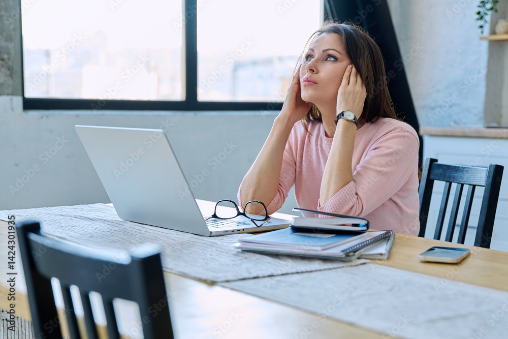 Middle aged sad stressed woman sitting at home at table with computer