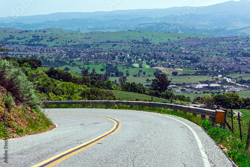 Scenic winding Quimby Road with guardrail curves downhill through green hills overlooking the expansive green landscape and suburban sprawl of Silicon Valley