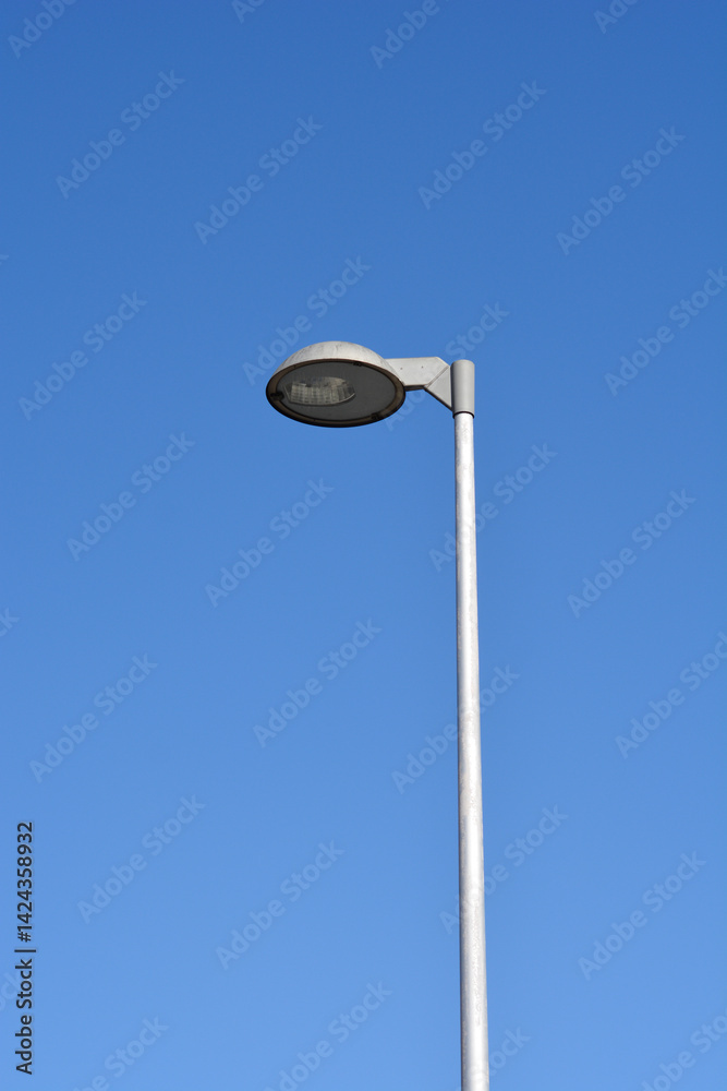 A minimalist close-up photograph of a street lamp with a round shade, mounted on a metal pole, against a solid blue sky