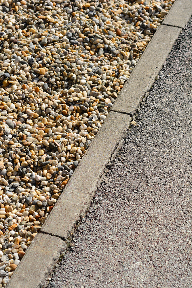 A close-up shows a gray concrete curb separating a patch of small, multicolored gravel from an asphalt surface