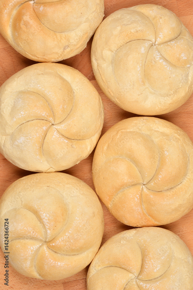 Kaiser rolls are arranged closely together on a light brown wooden surface, viewed from directly above