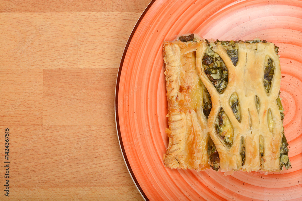 A slice of baked savory pastry with a lattice top and spinach filling sits on a colorful plate against a wooden surface