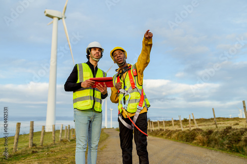 Obraz na płótnie Two engineers wearing safety vests and helmets are inspecting wind turbines, one holding a digital tablet and the other pointing towards a turbine w salonie