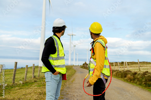 Obraz na płótnie Two engineers wearing safety vests and helmets are discussing next to wind turbines, showcasing renewable energy and teamwork w salonie