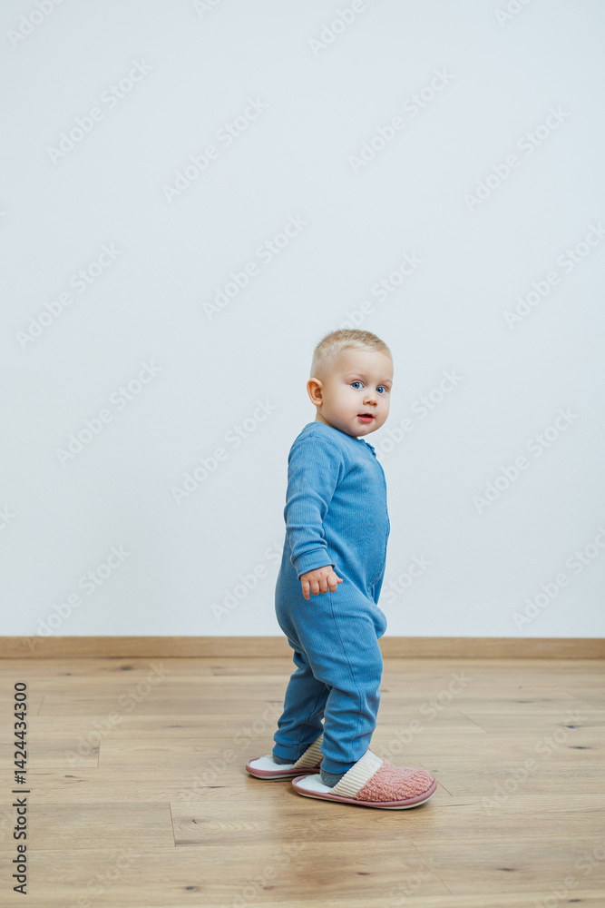 Little toddler boy in a blue romper tries on big house slippers. Child plays at home in the living room. Children's home clothes.