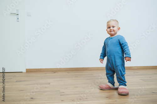 Obraz na płótnie Little toddler boy in a blue romper tries on big house slippers. Child plays at home in the living room. Children's home clothes. w salonie