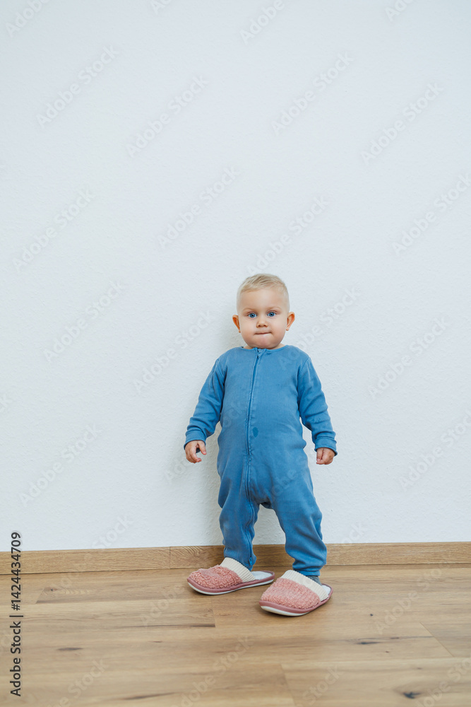 Little toddler boy in a blue romper tries on big house slippers. Child plays at home in the living room. Children's home clothes.