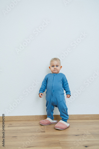 Little toddler boy in a blue romper tries on big house slippers. Child plays at home in the living room. Children's home clothes.