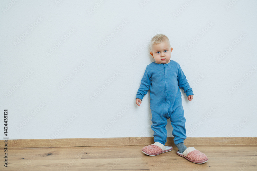 Little toddler boy in a blue romper tries on big house slippers. Child plays at home in the living room. Children's home clothes.